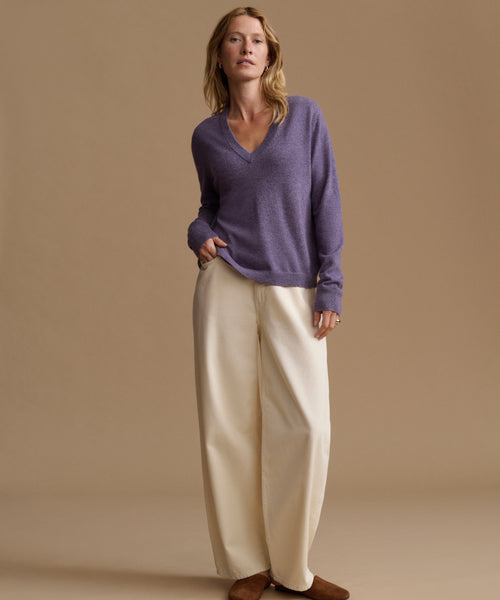 A woman stands against a plain beige studio background wearing a loose purple V-neck sweater, wide-leg cream pants, and brown shoes.