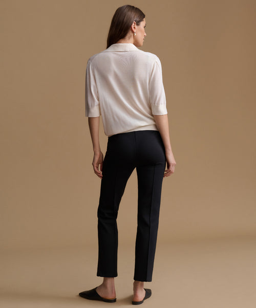 A woman shown from the back wearing a cream-colored knit polo shirt and black trousers, standing against a solid beige studio background.