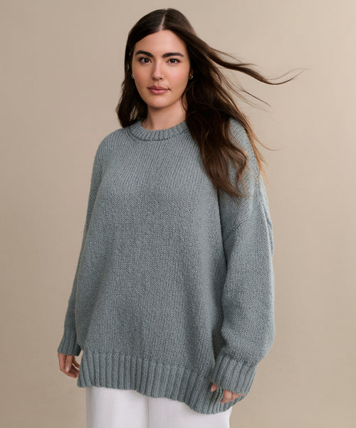 A woman with long brown hair stands against a beige background, her hair gently blowing to the side. She wears the oversized, light gray Foster Cotton Sweater with white pants.