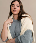 A woman with long brown hair wears the oversized Foster Cotton Sweater with a cream-colored sweater draped over her shoulders, standing against a neutral beige background. She gazes gently at the camera, exuding relaxed elegance.