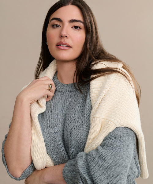 A woman with long brown hair wears the oversized Foster Cotton Sweater with a cream-colored sweater draped over her shoulders, standing against a neutral beige background. She gazes gently at the camera, exuding relaxed elegance.