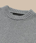 Close-up of the Foster Cotton Sweater, an oversized crewneck in light gray made from organic Tanguis cotton, featuring a ribbed neckline and black label inside the collar. Displayed against a plain beige background.
