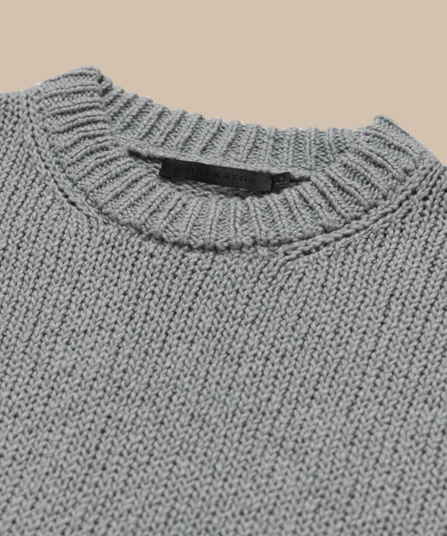 Close-up of the Foster Cotton Sweater, an oversized crewneck in light gray made from organic Tanguis cotton, featuring a ribbed neckline and black label inside the collar. Displayed against a plain beige background.