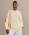 Wearing the oversized Foster Cotton Sweater in cream and white pants, a person stands against a beige backdrop, carrying a woven shoulder bag and smiling softly at the camera.