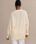 A person with short, dark hair is shown from behind wearing the Foster Cotton Sweater, an oversized crewneck in cream made from organic Tanguis cotton, paired with white pants, set against a plain beige background.