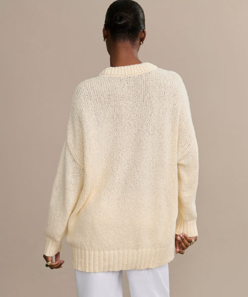 A person with short, dark hair is shown from behind wearing the Foster Cotton Sweater, an oversized crewneck in cream made from organic Tanguis cotton, paired with white pants, set against a plain beige background.