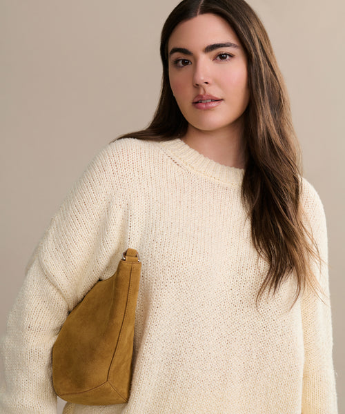 A woman with long brown hair wears the oversized Foster Cotton Sweater and carries a tan suede bag over her shoulder, standing against a beige background.