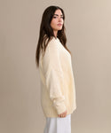A woman with long brown hair models the oversized Foster Cotton Sweater with white pants, standing in profile against a plain beige background.