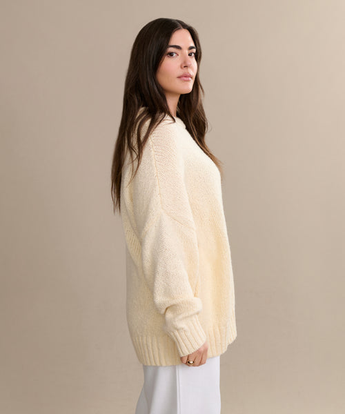 A woman with long brown hair models the oversized Foster Cotton Sweater with white pants, standing in profile against a plain beige background.