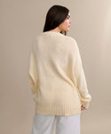 A woman with long dark hair, seen from behind, wears the oversized cream Foster Cotton Sweater and white pants, standing against a neutral beige background.