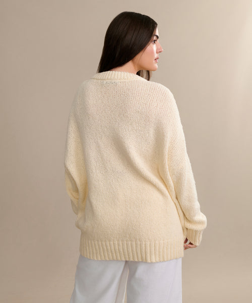 A woman with long dark hair, seen from behind, wears the oversized cream Foster Cotton Sweater and white pants, standing against a neutral beige background.