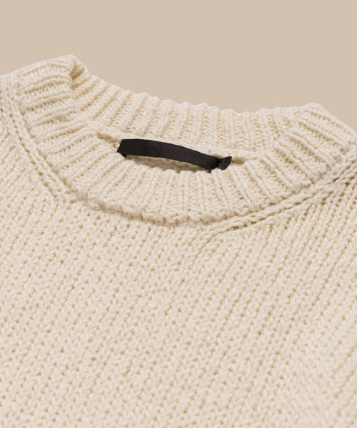 A close-up of the Foster Cotton Sweater, an off-white, chunky knit oversized crewneck with a ribbed neckline and black label inside the collar, made from organic Tanguis cotton and displayed against a beige background.
