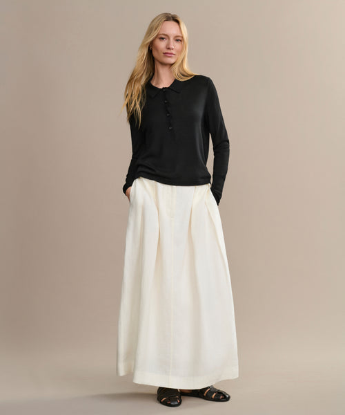 A woman with long blonde hair stands against a beige background wearing a black Grayson Pullover, a white ankle-length pleated skirt with pockets, and black sandals. Her hands rest in her skirt pockets.