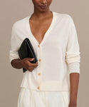 Wearing the Mira Cardigan in ivory with matching pants, a person holds a black clutch under their arm while standing against a neutral background.