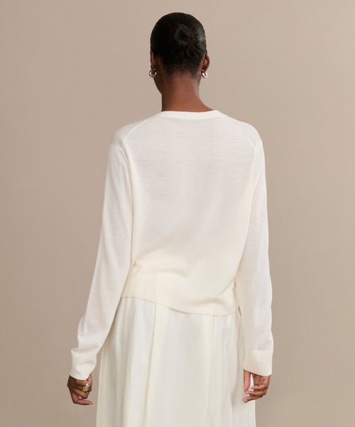 A person stands with their back to the camera, wearing the ivory Mira Cardigan and a flowing skirt. Their hair is styled in a neat bun against a plain beige background.