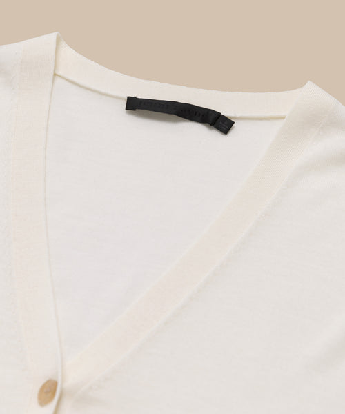 Close-up of the Mira Cardigan, a classic fit merino wool design in cream, with a black collar label and beige button on the left. Set against a neutral beige background.