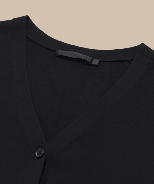 Close-up of the Mira Cardigan, a black knit V-neck cardigan with button detail and visible inner label, shown against a beige background.