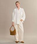 A woman poses against a neutral background in the boxy-fit white Linen Logan Shirt and wide-leg cream pants, holding a tan suede handbag. She wears matching flats with her hair pulled back, exuding relaxed and effortless style.