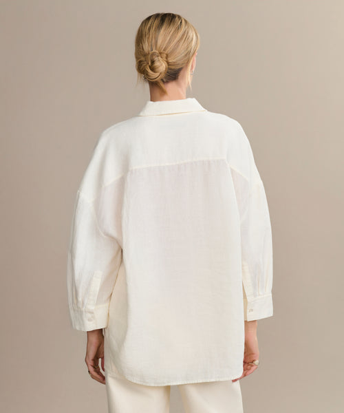 A woman with blonde hair in a low bun faces away, wearing the oversized, boxy-fit Linen Logan Shirt and matching white pants, embodying effortless style against a neutral background.