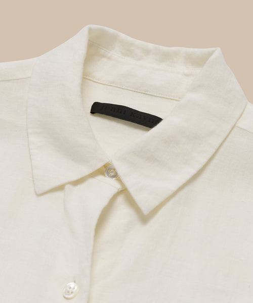 A close-up of the Linen Logan Shirt—a cream boxy-fit button-up with a pointed collar, white buttons, and a black label at the neck—laid flat on a neutral background.