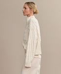 A woman with blonde hair in a low bun is shown in profile wearing the Cotton Cable Half Zip sweater and matching light-colored pants, standing against a plain beige background.