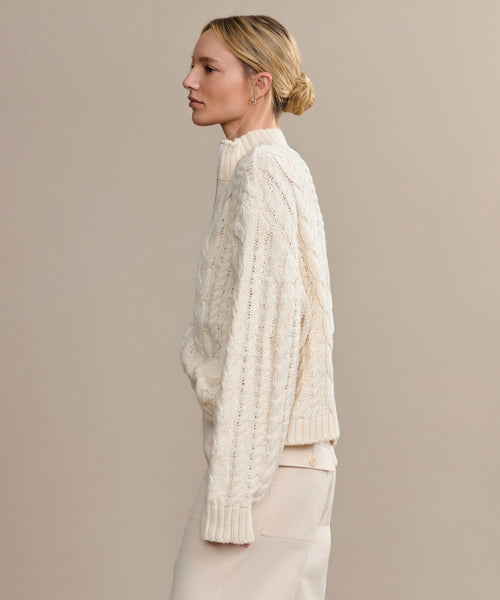 A woman with blonde hair in a low bun is shown in profile wearing the Cotton Cable Half Zip sweater and matching light-colored pants, standing against a plain beige background.