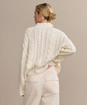 A woman with blond hair in a low bun faces away, wearing the Cotton Cable Half Zip sweater in ivory and high-waisted cream pants with flap pockets. Her left hand rests on her hip against a plain beige background.