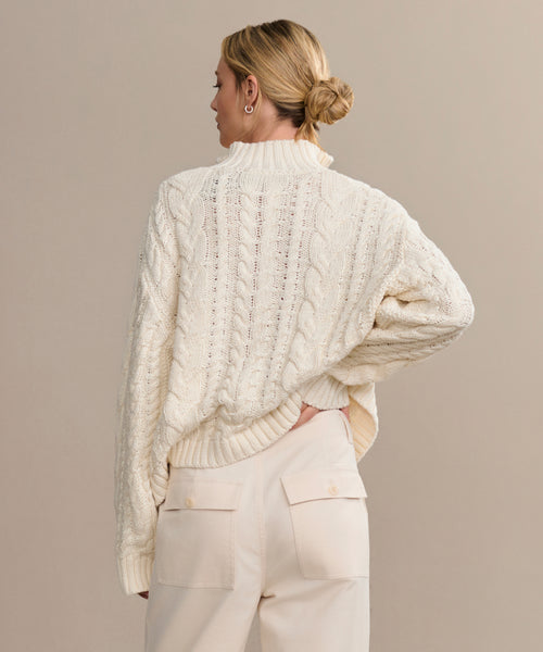 A woman with blond hair in a low bun faces away, wearing the Cotton Cable Half Zip sweater in ivory and high-waisted cream pants with flap pockets. Her left hand rests on her hip against a plain beige background.