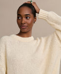 A woman wears the Foster Cotton Sweater, an oversized crewneck, looking to the side with a slight smile as she holds her hair back. The background is plain and neutral.
