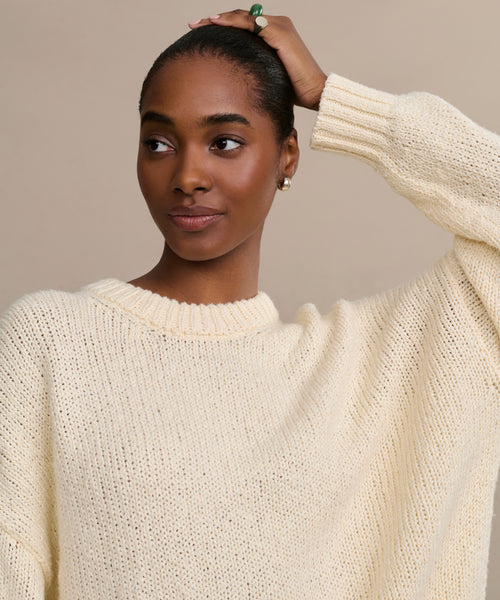 A woman wears the Foster Cotton Sweater, an oversized crewneck, looking to the side with a slight smile as she holds her hair back. The background is plain and neutral.