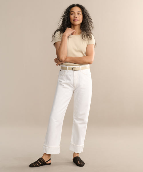 A woman with long curly hair stands confidently against a beige background, wearing a beige t-shirt, Fran Jean relaxed leg jeans with an untacked folded cuff, and black slip-on shoes with small gold accents.