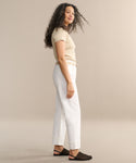 A woman with long curly hair stands sideways, smiling. She wears a beige short-sleeve top, relaxed leg jeans like the Fran Jean in Milkshake wash with an untacked folded cuff, and black woven slip-ons against a plain beige background.