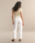 A person with long, curly hair faces away from the camera, wearing a beige short-sleeve shirt and Fran Jean relaxed leg jeans with an untacked folded cuff, paired with black sandals in a neutral studio setting.
