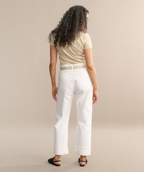 A person with long, curly hair faces away from the camera, wearing a beige short-sleeve shirt and Fran Jean relaxed leg jeans with an untacked folded cuff, paired with black sandals in a neutral studio setting.