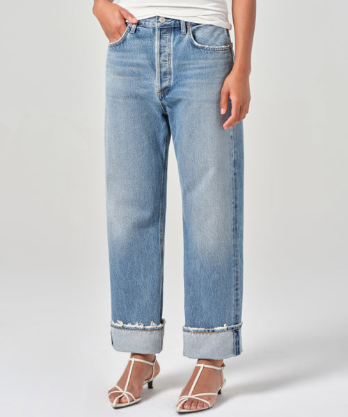 A person models the high-waisted, light blue Fran Jean with relaxed legs and wide, untacked folded cuffs, paired with a white top and strappy sandals, standing against a plain light background.