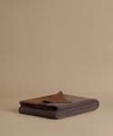 A folded dark gray wool blanket with a contrasting brown color on top, displayed on a warm beige studio background.