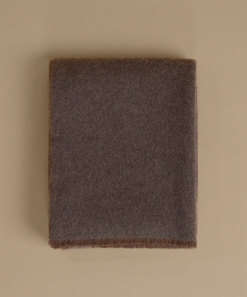 A folded dark brown woolen blanket with fringed edges, displayed flat against a solid beige background.