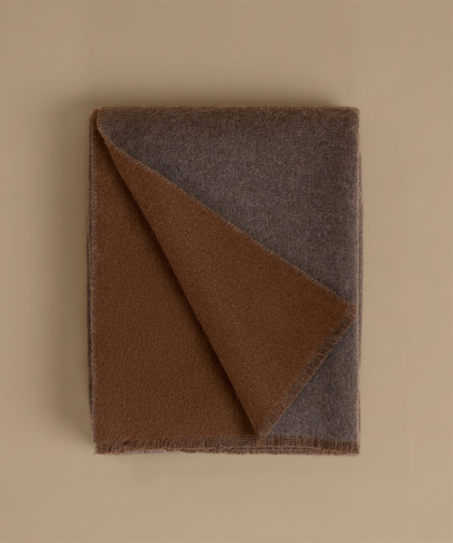 A folded wool blanket with two distinct colors: a warm brown side and a muted gray-brown side, displayed on a solid beige background.