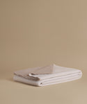 A folded stack of soft woven textiles in warm off-white and light beige tones, photographed on a plain beige studio background.