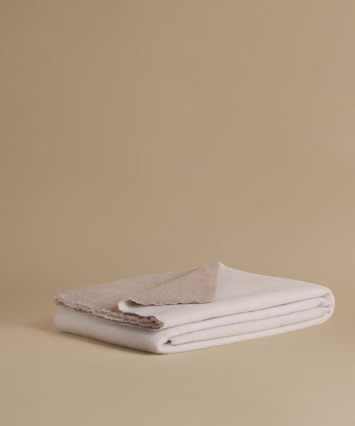 A folded stack of soft woven textiles in warm off-white and light beige tones, photographed on a plain beige studio background.