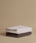 Two folded blankets, one in off-white and one in dark brown, stacked on a warm beige background.