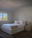 A bedroom featuring a low-profile platform bed with white bedding and beige accent pillows, flanked by two light wood nightstands with matching table lamps, set against a neutral cream-colored wall and ceiling.