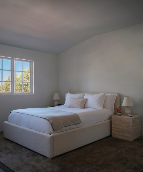 A bedroom featuring a low-profile platform bed with white bedding and beige accent pillows, flanked by two light wood nightstands with matching table lamps, set against a neutral cream-colored wall and ceiling.