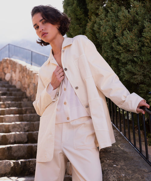 A person with short, dark hair wears the cream-colored Gibson Jacket and matching pants, standing outdoors near stone steps and greenery, one hand resting on a black railing.