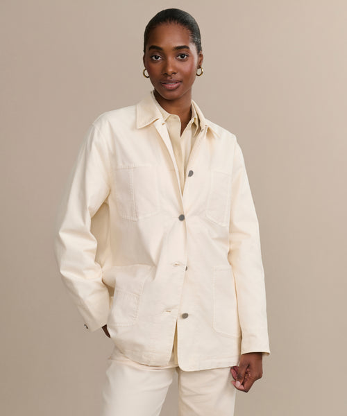 A woman stands against a beige background, wearing the cream-colored Gibson Jacket with large front pockets and matching pants. Her hair is pulled back as she looks at the camera, one hand by her side and the other touching her jacket.
