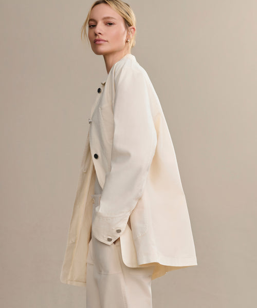 A woman with blonde hair stands sideways against a plain beige background, wearing the Gibson Jacket—a loose, off-white, summer-weight style—paired with matching pants.