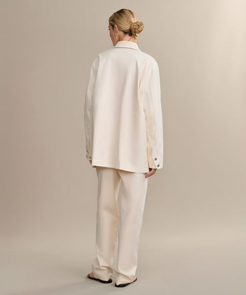 A person faces away, wearing the loose-fitting Gibson Jacket in off-white with matching pants. Their hair is styled in a low bun, and they stand barefoot against a neutral beige background.