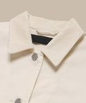 Close-up of the Gibson Jacket, a cream-colored summer-weight style featuring two silver snap buttons and a black label inside the collar, shown against a beige background.