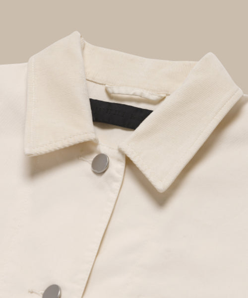 Close-up of the Gibson Jacket, a cream-colored summer-weight style featuring two silver snap buttons and a black label inside the collar, shown against a beige background.