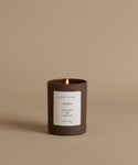 A lit candle in a matte dark brown glass jar with a white label reading 'jenni kayne' and 'Amber', set against a solid warm beige background.